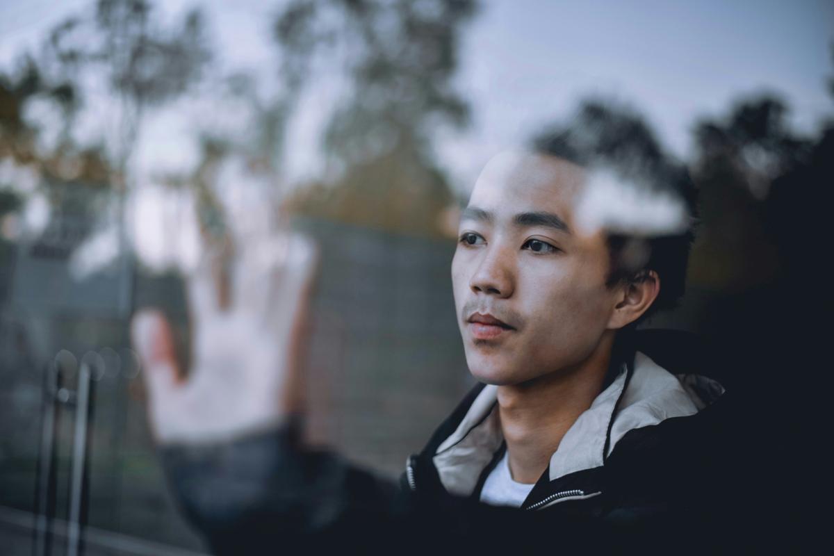 Man looking through a window with hand raised, symbolising low self-confidence, self-doubt, and emotional distance explored in psychodynamic therapy.