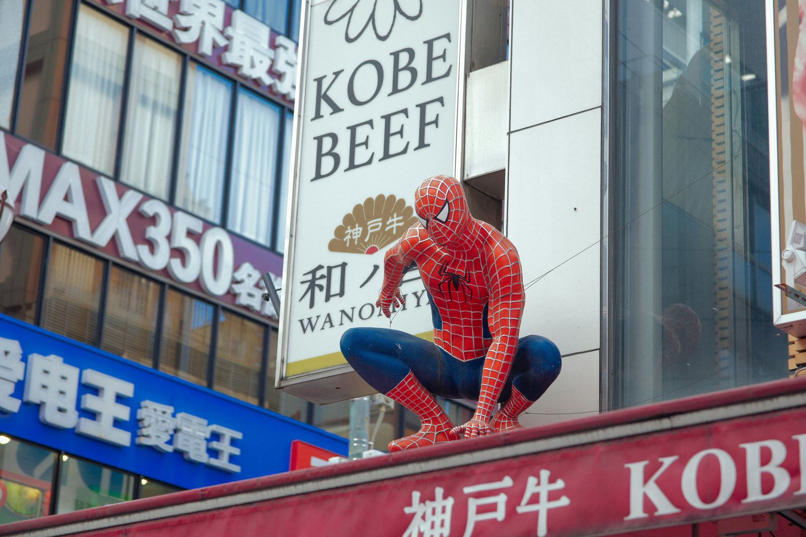Spider-Man figure crouching above a Kobe Beef shopfront sign, symbolising the weight beneath a hero's confident exterior
